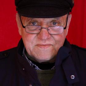 Man in black coat, hat and glasses in front of a red curtain