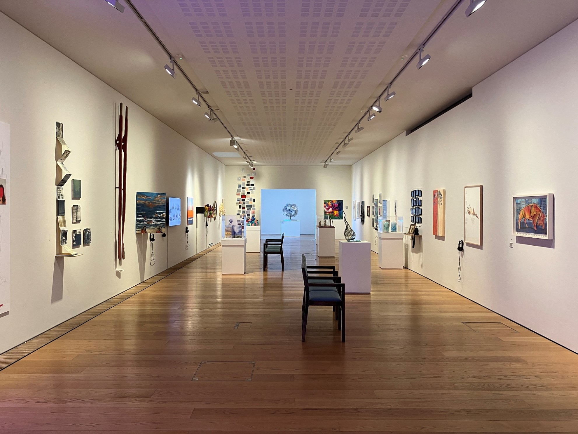 A long room with artwork on all the walls. In the middle of the room are two dark wooden benches and 5 white plinths that have sculptures on them.