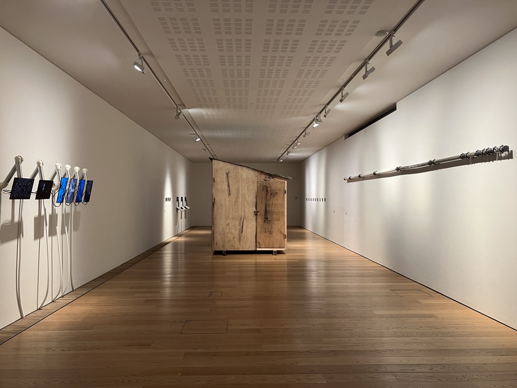 a long room with white walls and a shiny wooden floor. on the left hand wall here are 5 screens protruding from the wall. In the middle of the room is a wooden shed with a slanted roof. On the right wall is a long tube suspended the length of the wall.