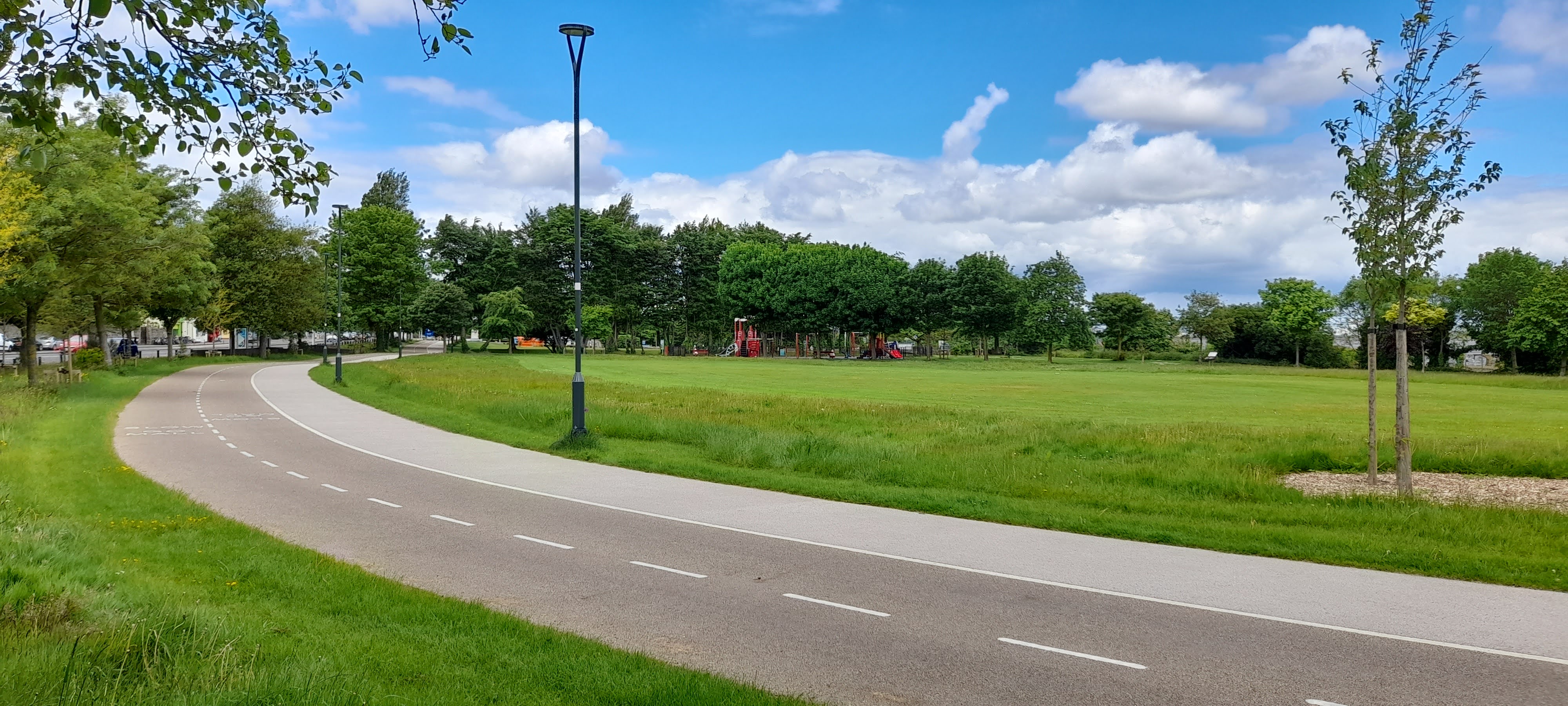 A curved, paved path bordered by grass and trees extends through a park under a blue sky with clouds. A playground with red equipment is visible in the distance.