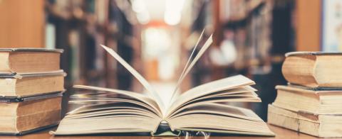 An open book in the centre of the image with other books stacked beside it, library corridor out of focus in the background